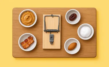 Illustration: top-down flat lay on a wooden board: a rat snap trap in the center surrounded by small bowls of bait options: peanut butter, a dried fruit piece, a bacon bit, a nut, a cotton ball.