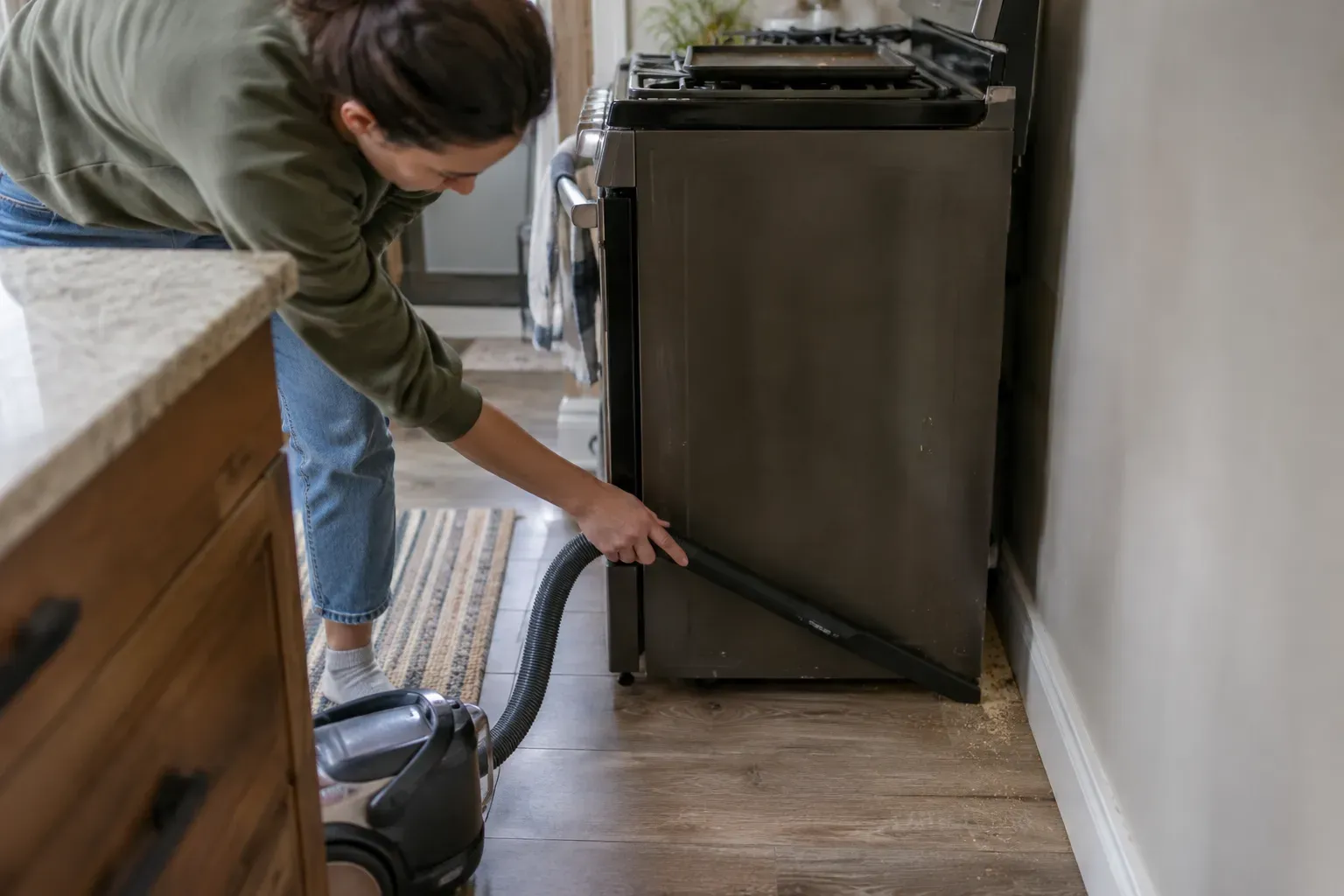 A person vacuuming crumbs from the gap beside a pulled-out kitchen stove with a crevice tool